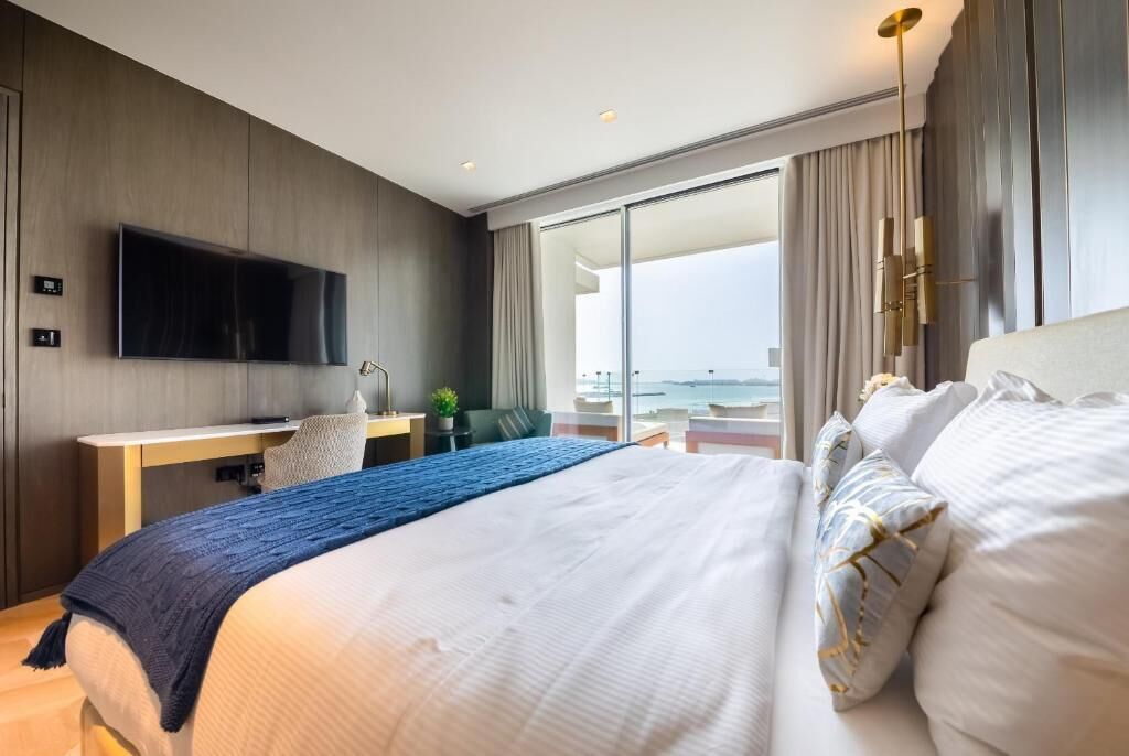 A modern hotel room with a large bed, a desk with a flat-screen TV, a chair, and a view of the sea through large windows.
