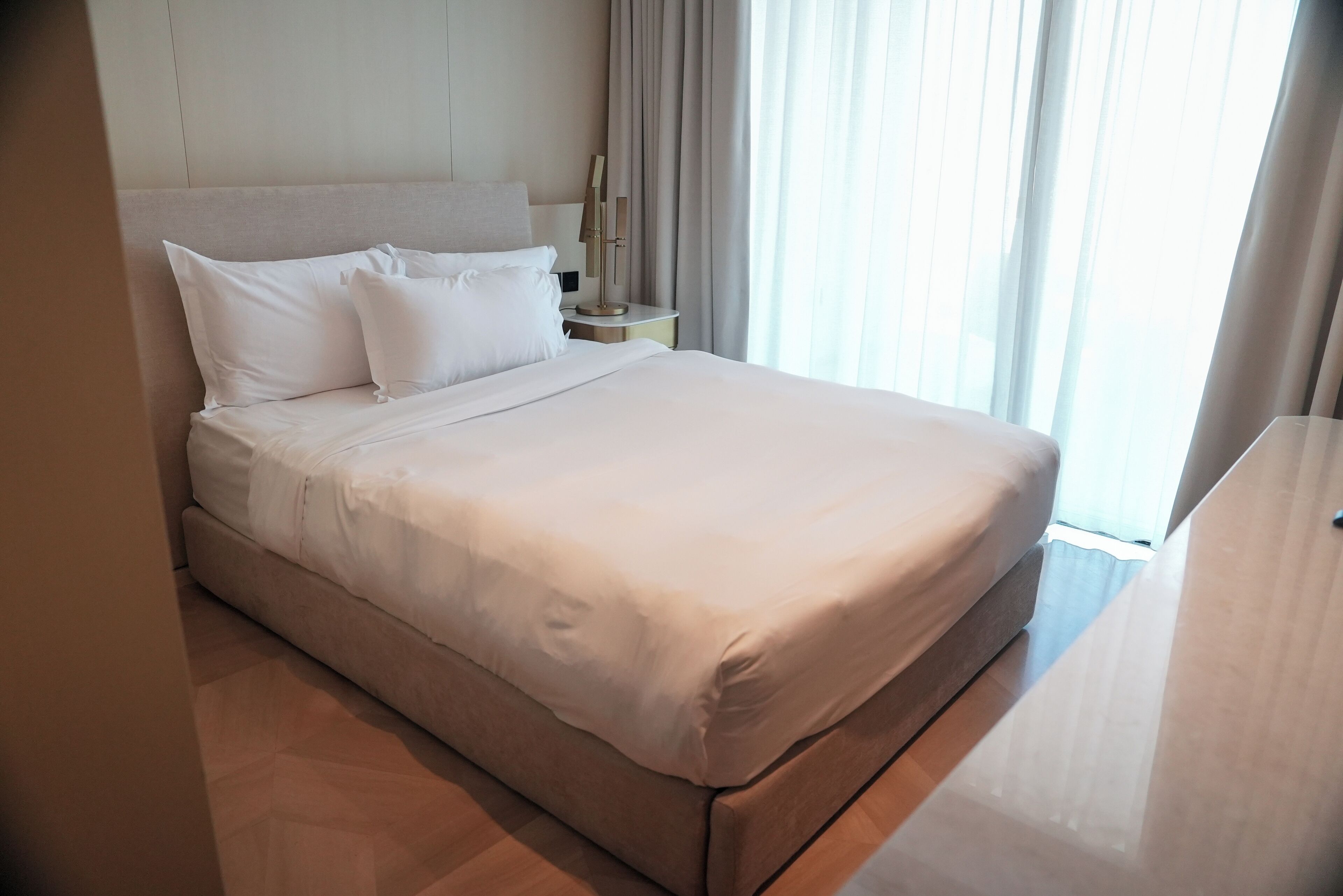 Una cama bien hecha con sábanas blancas, una mesita de noche con lámpara y una ventana con cortinas.