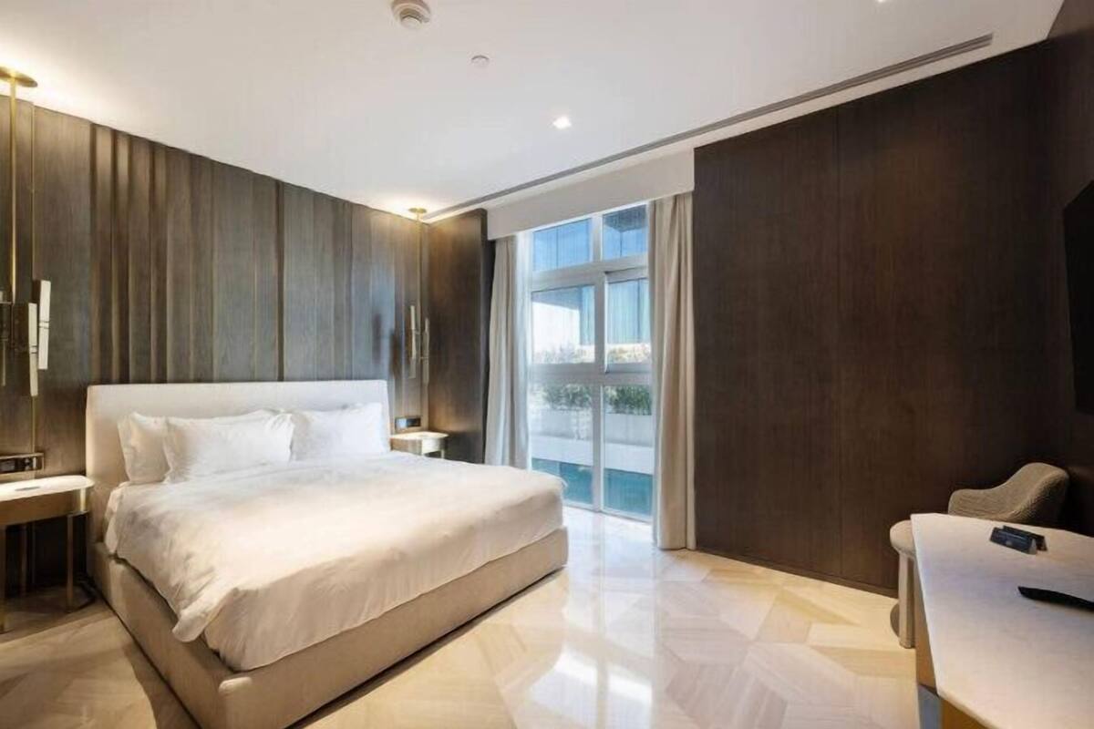 A modern hotel room with a large bed, a desk, and a view of the outdoors through a large window.