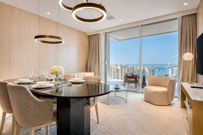 A modern hotel room with a dining table set for four, a sofa, a chair, and a view of the cityscape.
