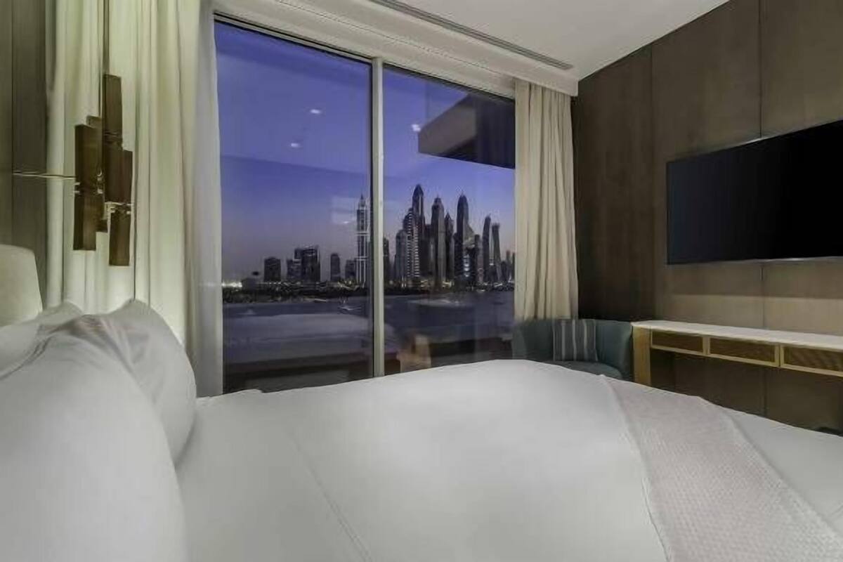 A hotel room with a large bed, a TV mounted on the wall, and a view of a city skyline through a large window.