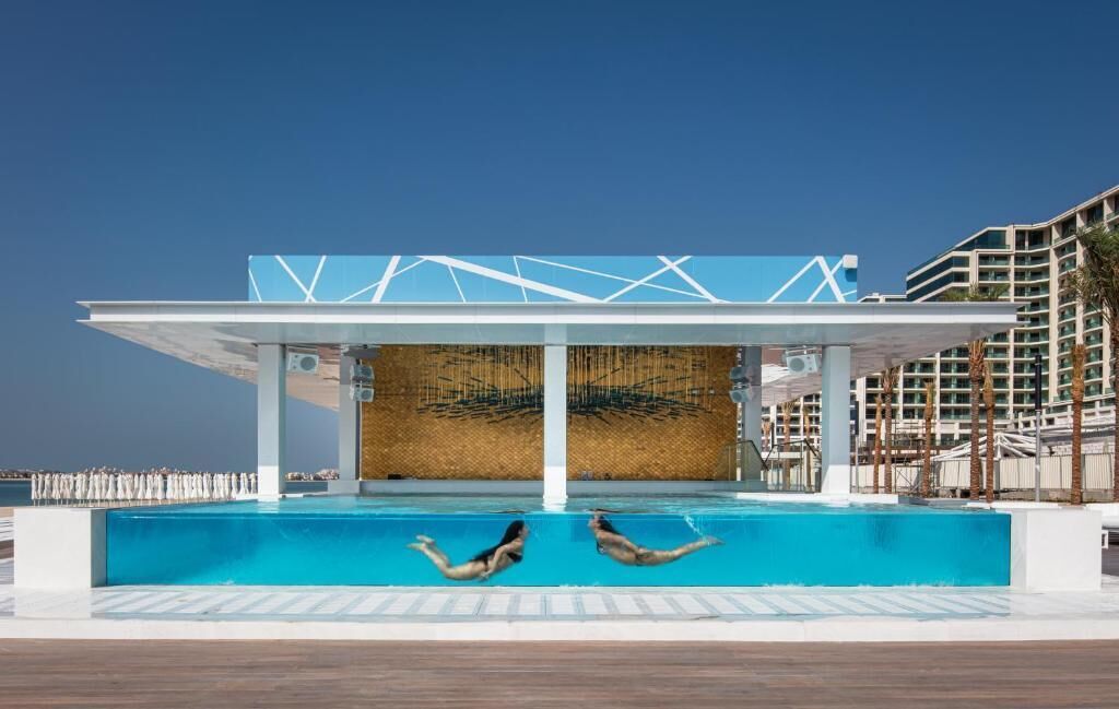 Una piscina con due persone che si tuffano, una zona ombreggiata con motivi geometrici e un edificio con balconi sullo sfondo.