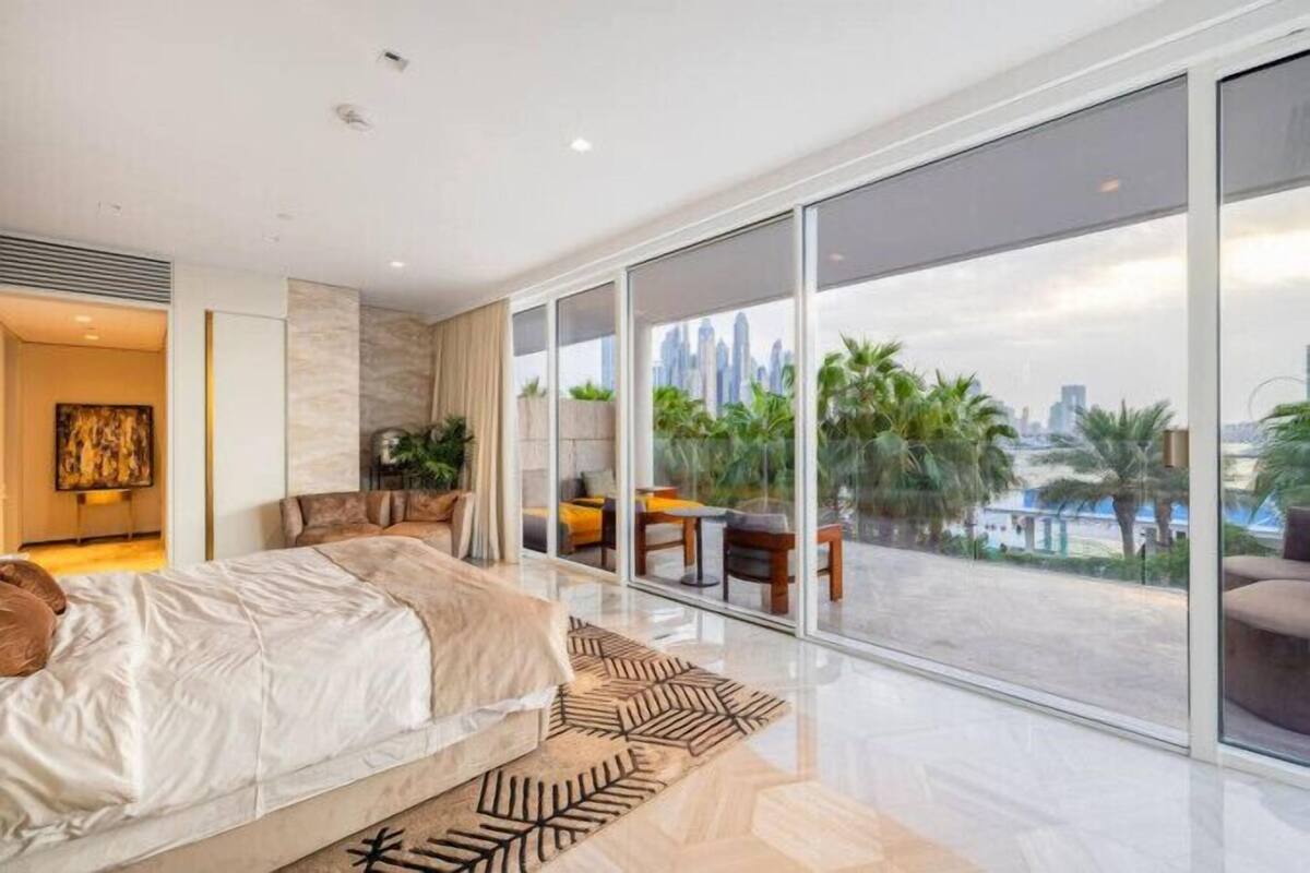 A modern bedroom with a large bed, a view of the cityscape through floor-to-ceiling windows, and a balcony with outdoor furniture.