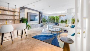 Een moderne woonkamer met een bank, salontafel en barkrukken. Er is een grote televisie aan de muur bevestigd, en de kamer heeft een groot raam met uitzicht op groen.