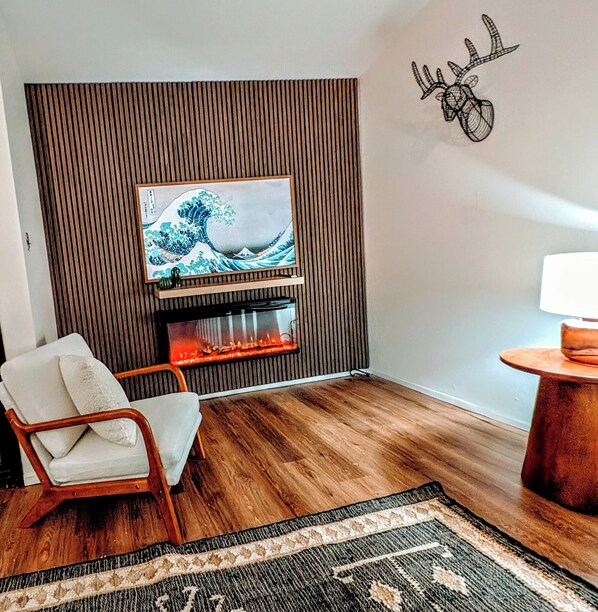 Een woonkamer met een flatscreen televisie bevestigd aan een houten paneelwand, een open haard, een witte fauteuil, een vloerkleed met patroon en een kleine ronde tafel met een lamp.