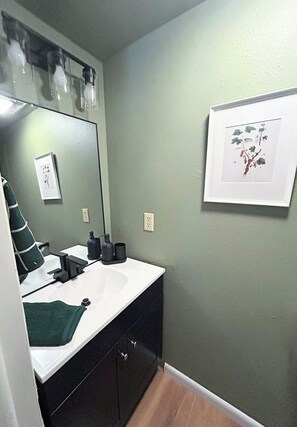 Een badkamer met een wit wastafelmeubel, donkere kastjes, een groene handdoek en een lijst met een botanische afbeelding aan de muur.