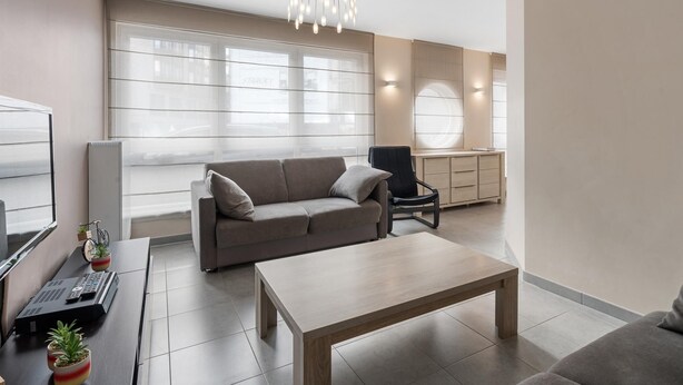 Un salon moderne avec un canapé, une table basse et un téléviseur. On y trouve un bureau avec un ordinateur et une chaise. La pièce est dotée de grandes fenêtres et d’un luminaire au plafond.