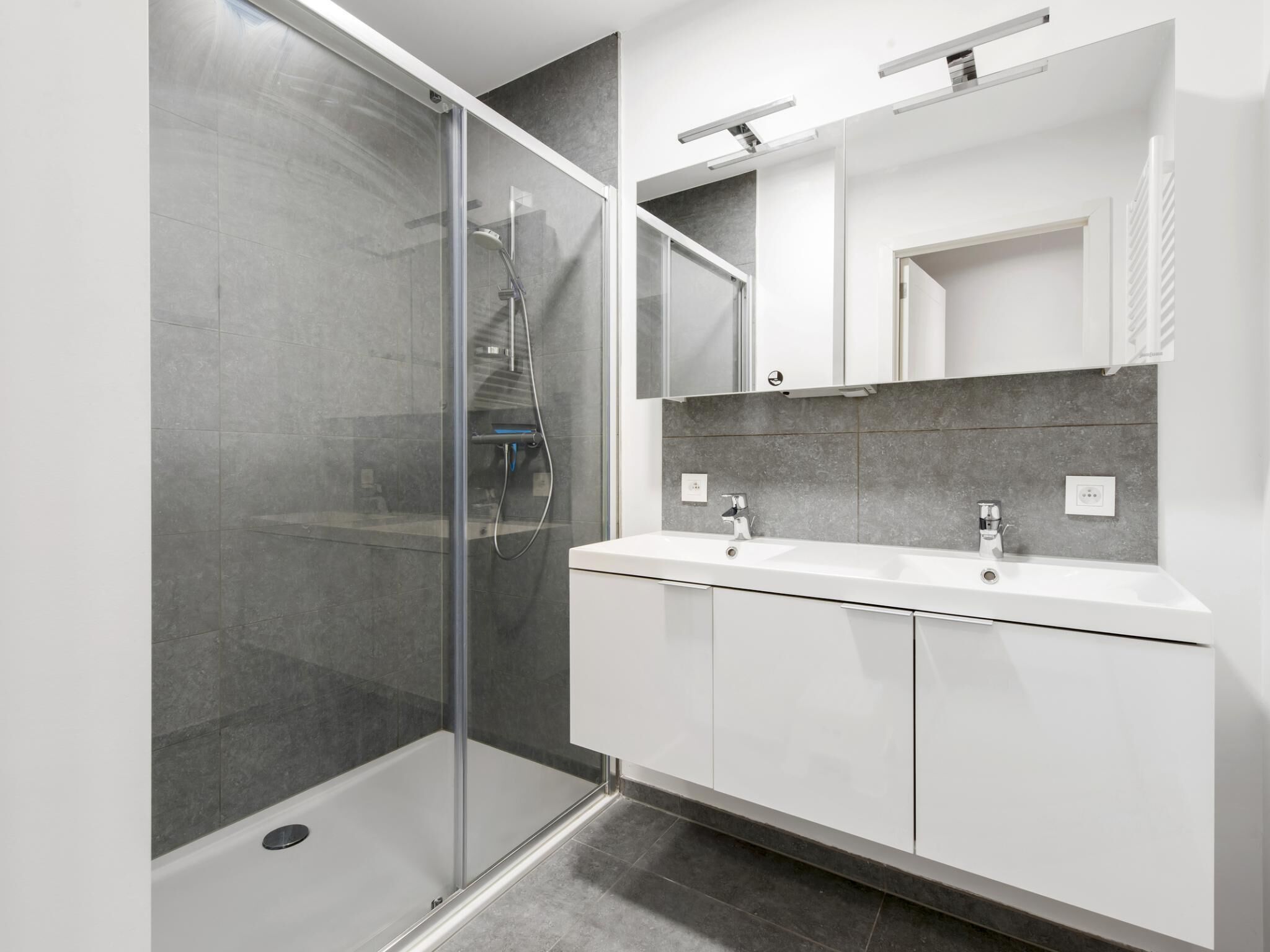 A modern bathroom with a glass-enclosed shower, a white vanity with two sinks, and a large mirror above the vanity.