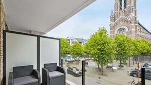 Un balcon avec des meubles en osier, une vue sur une église et une rue bordée d’arbres et où circulent des voitures.