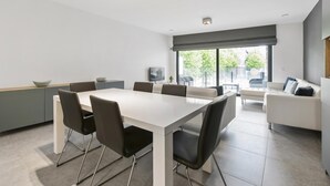 Un espace repas moderne avec une table blanche et des chaises noires, un canapé gris et une grande fenêtre donnant sur un paysage verdoyant.