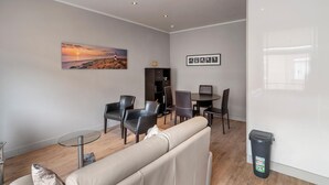 A modern living room with a beige sofa, a glass coffee table, and a dining area with a dark wooden table and chairs. There is a painting of a sunset on the wall, a bookshelf, and a trash bin. - Ideal holiday home with garage (Blankenberge)