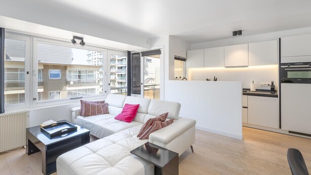 A modern apartment with a living area featuring a white sofa, a coffee table, and a view of the city through large windows. - Modern apartment near the sea (Middelkerke)
