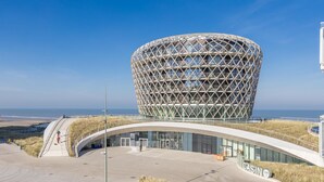 Un edificio moderno con una fachada geométrica distintiva, una entrada curva y vista al mar de fondo.