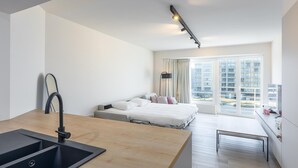 Una habitación de hotel moderna con kitchenette, una cama con muchas almohadas, un ventanal con vistas a edificios y un escritorio con silla.