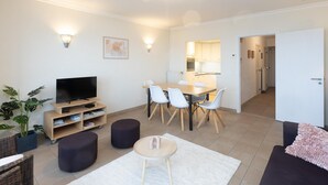 Un salon moderne avec un téléviseur à écran plat, une table à manger avec des chaises, un canapé et une petite table basse. On aperçoit un coin cuisine à travers une porte ouverte.