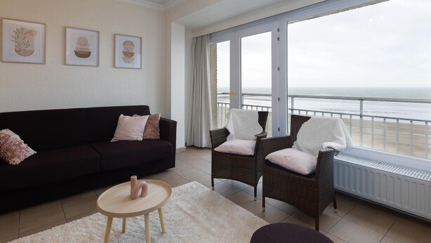 Un salon avec un canapé aux couleurs sombres, deux fauteuils en osier, une petite table ronde et une vue sur la mer grâce à de grandes portes vitrées.