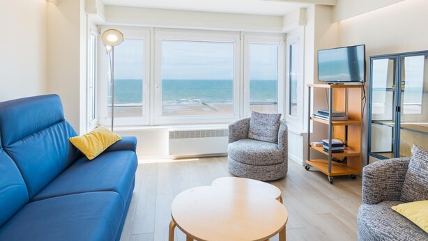 Un salon moderne avec un canapé bleu, un fauteuil gris et une table basse ronde. Un téléviseur est fixé sur un meuble en bois, et une grande fenêtre offre une vue sur la mer.