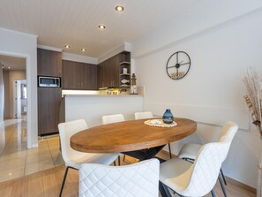 Ein moderner Essbereich mit einem ovalen Holztisch, weißen Polsterstühlen, einer Küche im Hintergrund und einer Wanduhr.