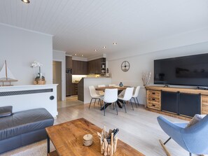 Ein modernes Wohnzimmer mit einer Couch, einem Holztisch und einem Fernseher. Es gibt einen Essbereich mit Tisch und Stühlen. Im Hintergrund ist eine Küche zu sehen.