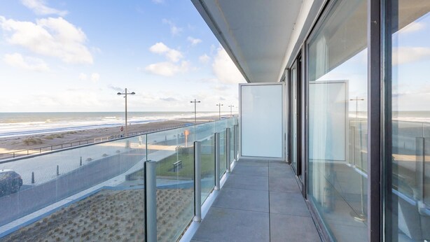 Een balkon met glazen hekwerk met uitzicht op een strand en een weg.