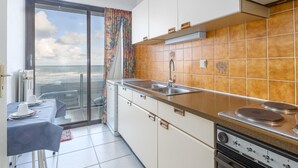 Een compacte keuken met uitzicht op zee, een eethoek met een tafel voor twee en een glazen schuifdeur naar een balkon.