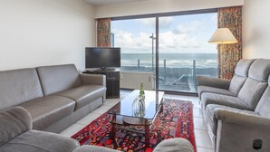 Een moderne woonkamer met een bank, fauteuils, een televisie en uitzicht op de oceaan door grote ramen.