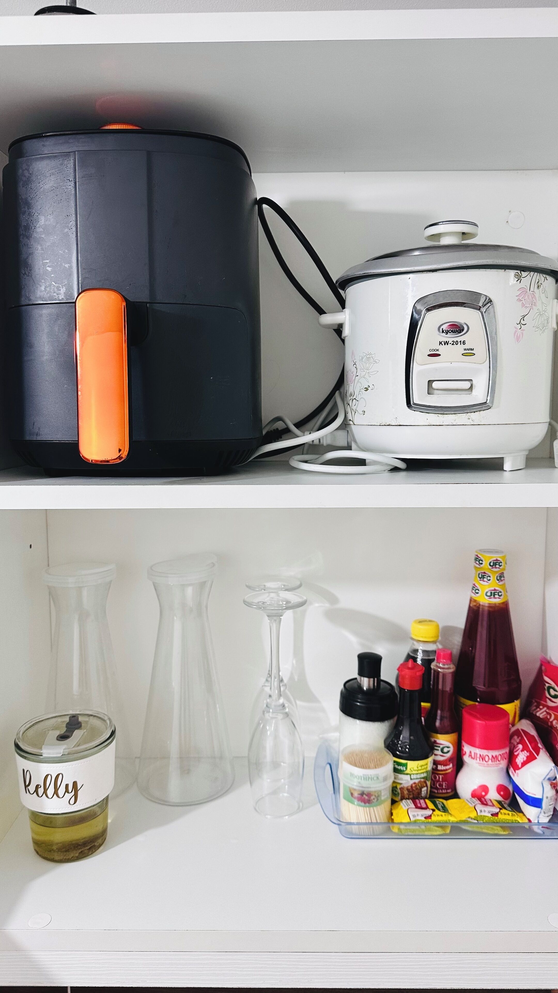 A kitchen with a black appliance and a white rice cooker on the countertop. Below, there are various kitchen items including a jar labeled "Kelly," a wine glass, and several condiment bottles.