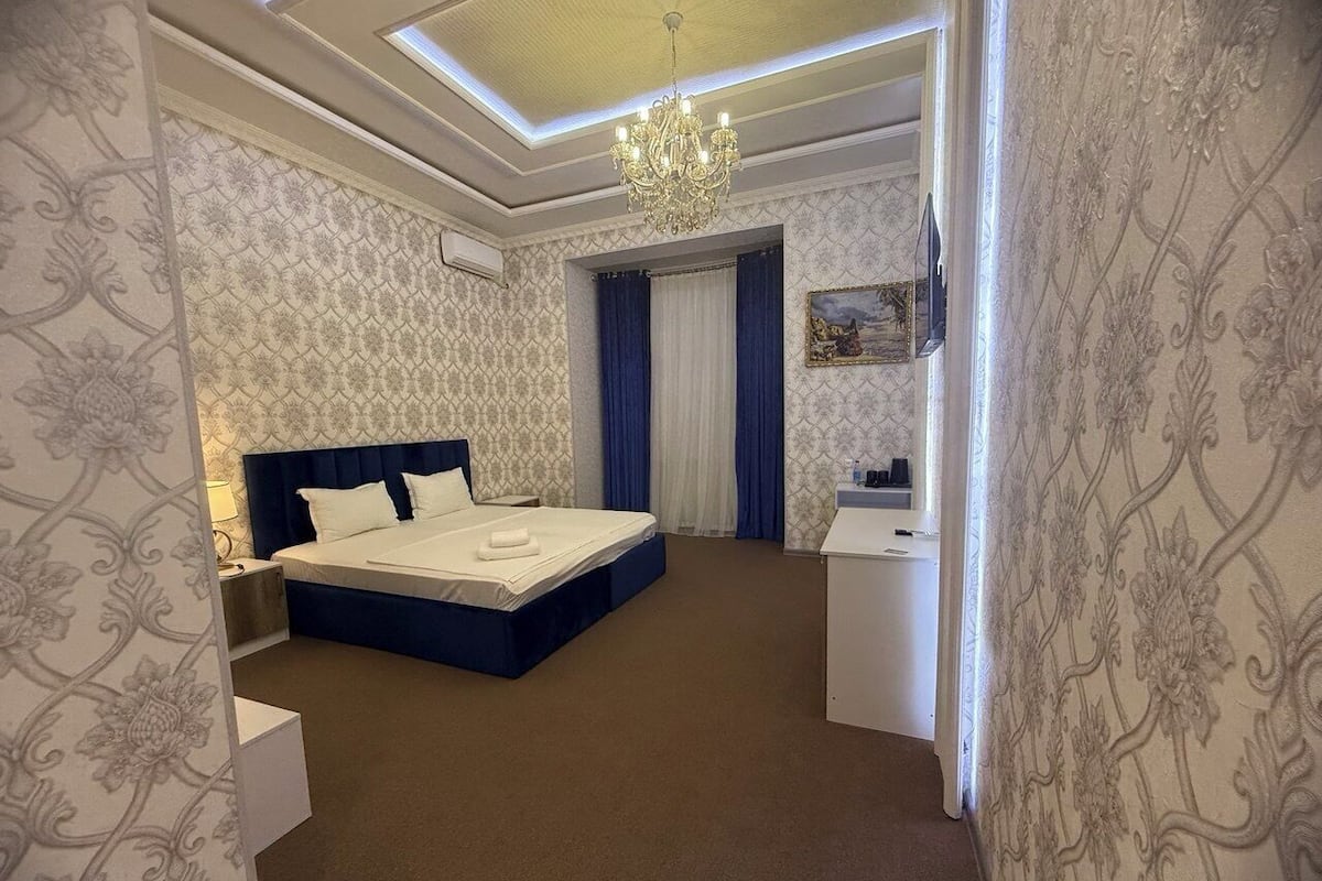 A hotel room with a bed, bedside table, a desk, a chair, a chandelier, and patterned wallpaper.
