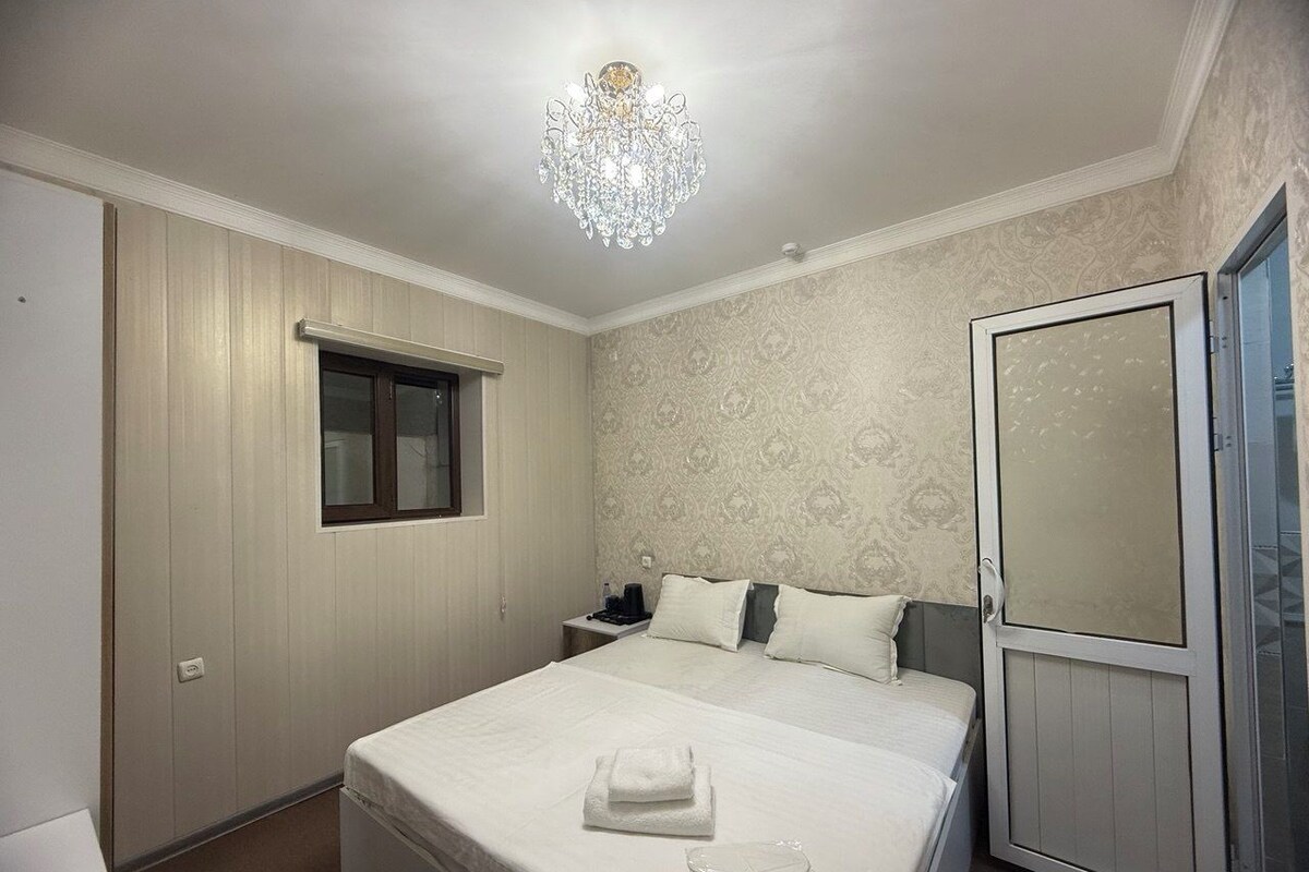 A bedroom with a bed, a nightstand, a window, a chandelier, and a wardrobe.