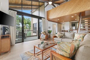 A spacious living area with a large sliding glass door leading to a balcony, a wooden ceiling, and a kitchen visible through an open door. - Oceanfront Complex - 2bdrm/2bath with Loft - NEW! Sleeps up to 8! AC, Wifi, more (Kailua-Kona)