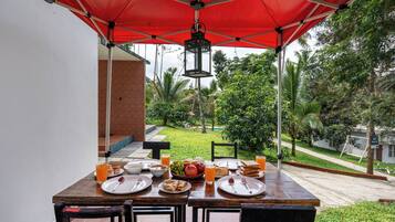 An outdoor dining area with a red umbrella, a table set with plates, glasses, and a bowl of fruit.