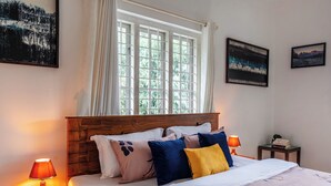 A neatly made bed with a floral bedspread, a wooden headboard, and multiple pillows. There are two bedside lamps, a small table with a book, and a window with curtains. - Exuberance Stays (Sulthan Bathery)