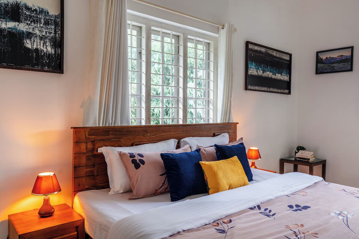 A neatly made bed with a floral bedspread, a wooden headboard, and multiple pillows. There are two bedside lamps, a small table with a book, and a window with curtains.