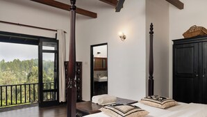 Une chambre à coucher avec un grand lit, un lit à baldaquin en bois, une table de chevet, un miroir, une armoire et un balcon avec vue sur les arbres.