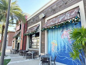 An outdoor seating area with tables and chairs, a mural on the wall, and palm trees. - Margaritaville Luxe Cottage • 2 Master Suites • Disney ~5m • Walk to Fun & Eats (Kissimmee)