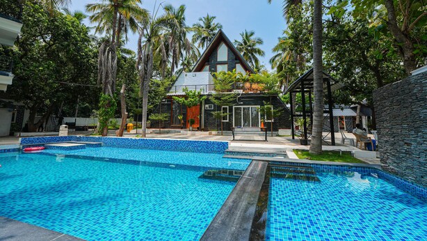 A pool area with clear blue tiles, surrounded by palm trees and a building with a triangular roof. - MOON LIGHT RESORT (Idappadi)