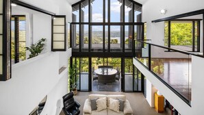 Un interior modern amb un sostre alt, grans finestres que ofereixen vistes a l'exterior, una zona de dinar amb taula rodona i una disposició de seients amb sofà i cadires.