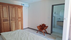 Ein Schlafzimmer mit einem Bett, einem Holzschrank und einem Sessel. Ein Badezimmer ist durch eine offene Tür sichtbar.