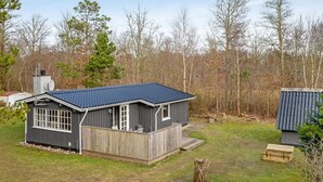 Een gelijkvloers huis met een donker dak en grijze buitenbekleding, omgeven door een houten terras en een omheinde tuin.