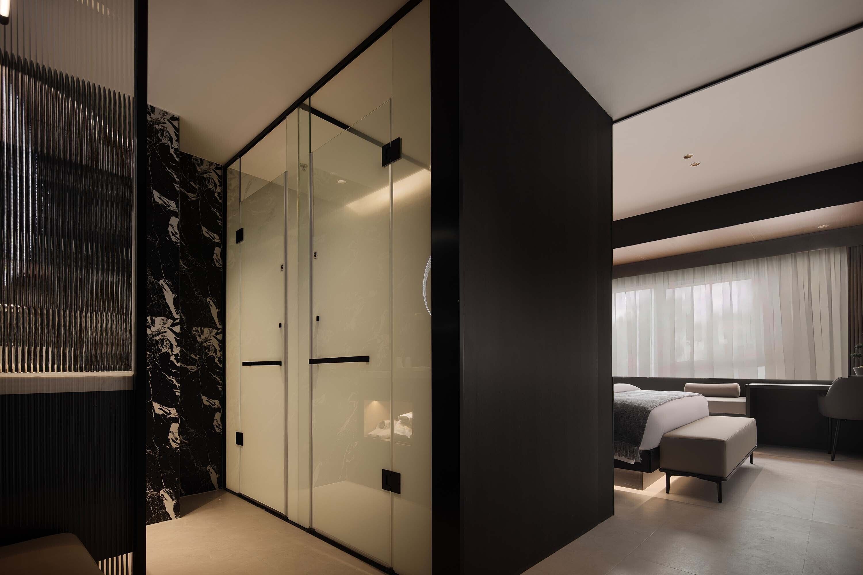 A modern hotel room with a glass partition, a bed with a headboard, a seating area, and a large window with curtains.