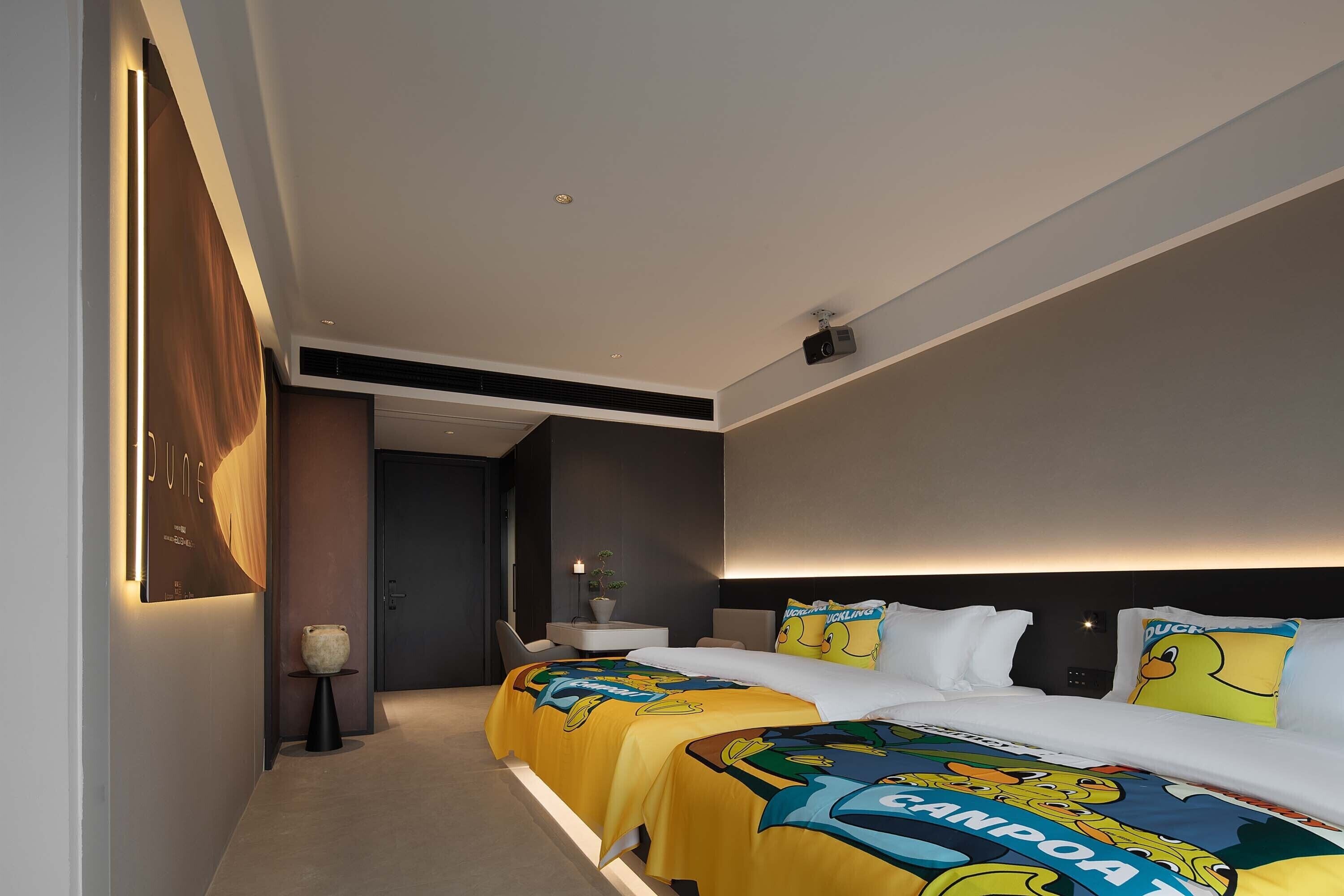 A hotel room with two beds, a large poster on the wall, a small table with a vase, and a night light.