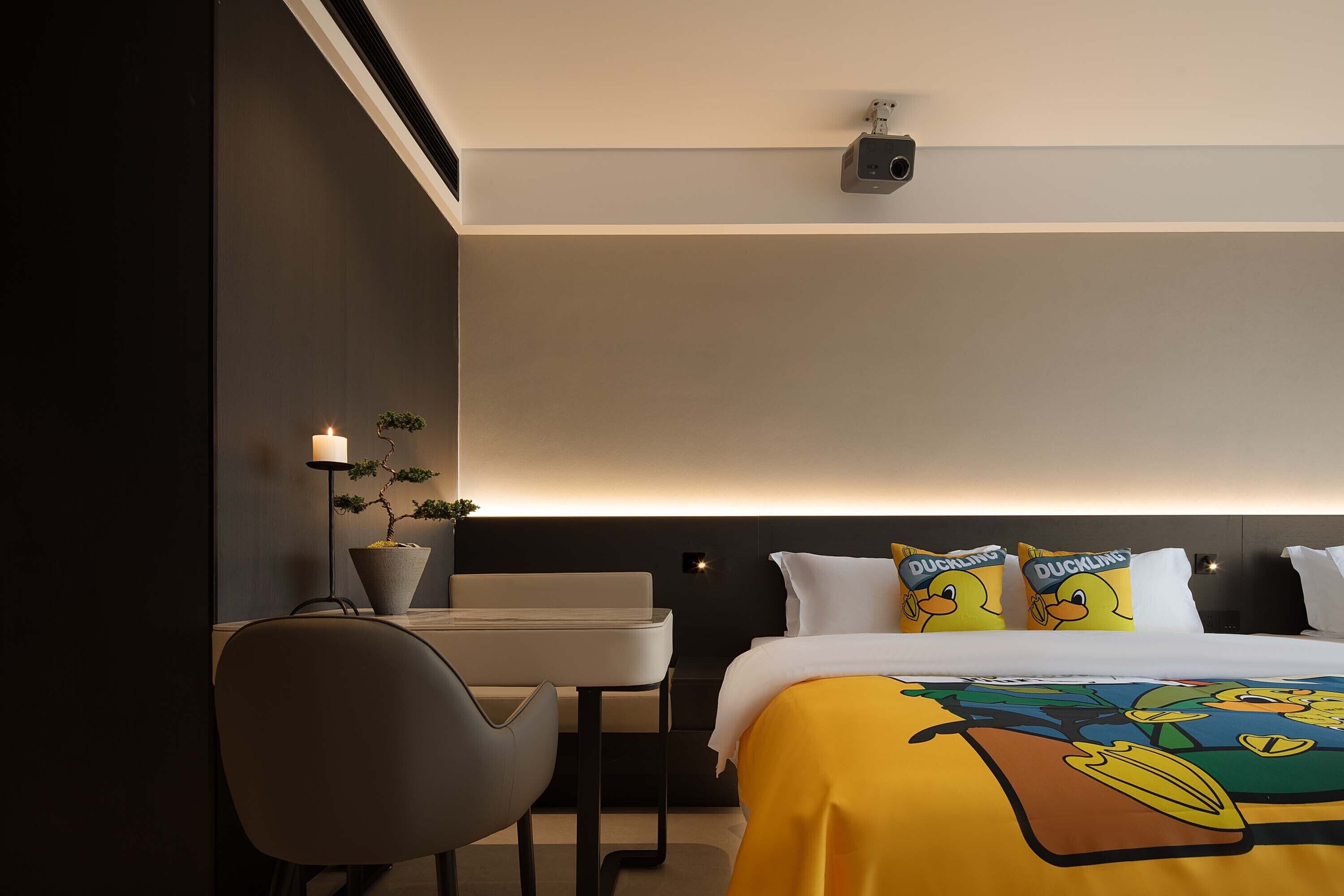 A hotel room with a bed, a desk, a chair, a potted plant, and a wall-mounted projector.