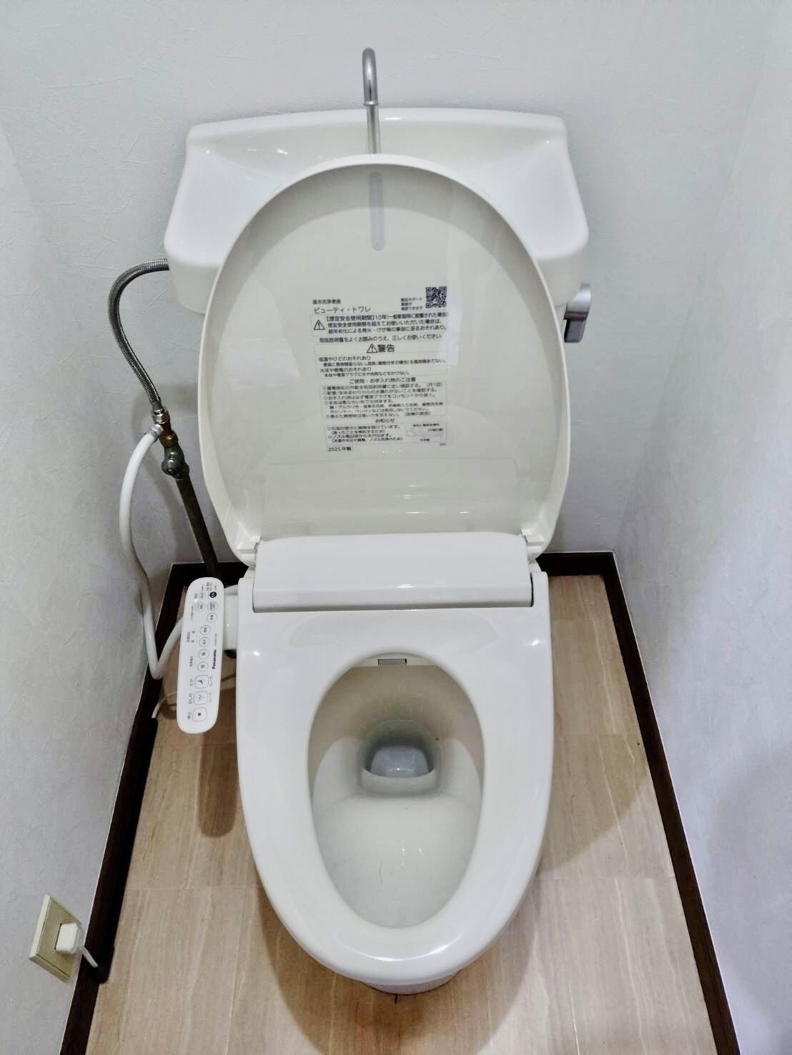 Uma sanita branca com painel de comando à esquerda, um código QR na tampa e uma etiqueta de aviso.