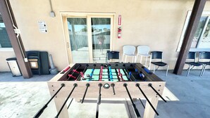 Un tavolo da calcio all'aperto con un estintore nelle vicinanze, un cestino e due sedie appoggiate a una parete con una porta e un cartello.