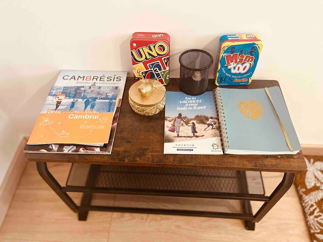 Una mesa de madera con un libro titulado 'Cambrai', una caja de cartas UNO, una caja de M&M's, un recipiente pequeño de malla negra y una libreta con diseño de hoja tropical.