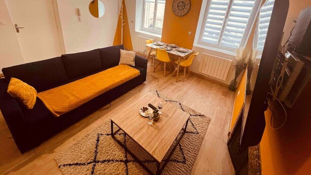 A living room with a black sofa, a wooden coffee table, and a dining area with yellow chairs. - Le Ch`ti Cambrai, Logement Élégant, Centre ville (Cambrai)
