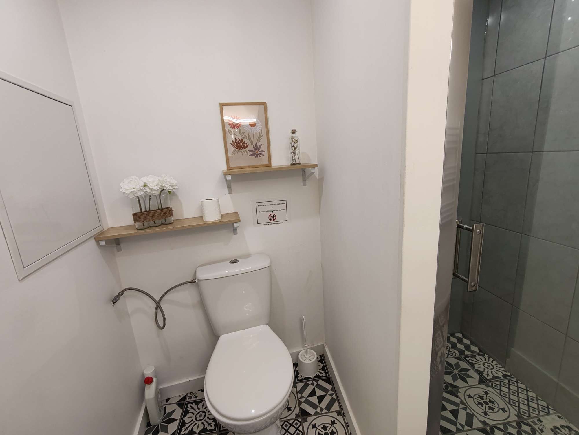 A small bathroom with a toilet, a shower, and a sink. There are decorative items on the wall and patterned tiles on the floor.
