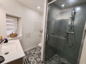 Un baño con ducha de vidrio, lavamanos blanco, pisos de baldosas estampadas, una ventana con persianas y una cesta en el alféizar.