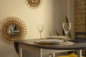 Una mesa de comedor con dos copas de vino, platos y cubiertos, frente a una pared con un espejo decorativo y una planta.