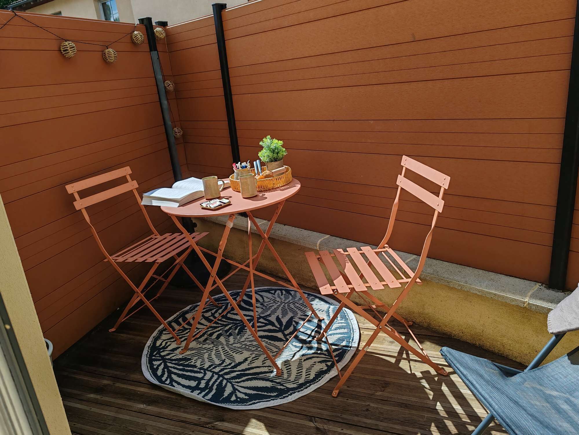 A small outdoor seating area with a round table, two chairs, a potted plant, and a patterned rug.